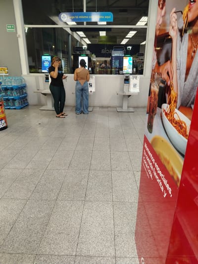 Here is a description of the image:
**Scene:** An interior shot of a store self-checkout area, likely in Tinaquillo, Venezuela based on the Spanish signage.
**People:** Two women are present. One is wearing a black top and dark leggings and is on a cell phone. The second woman is wearing an off-the-shoulder top and low-rise, wide-leg jeans.
**Objects:**
* Several self-checkout kiosks are visible.
* Stacks of bottled water are on display.
* A promotional stand featuring an image of a person eating pasta and drinking Coca-Cola is in the foreground.
* Shopping bags are visible on the kiosks.
**Environment:** The scene is well-lit with tiled flooring. Signage is in Spanish. The atmosphere appears to be a typical supermarket or department store setting.