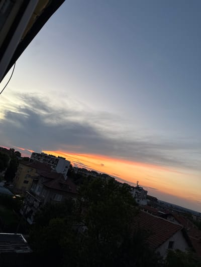 The image captures a sunset over a city. The sky is a gradient of blue and gray, with streaks of orange and pink clouds near the horizon. Below the sky, there is a cityscape with various buildings, some appearing to be apartment blocks and others smaller residential houses with tiled roofs. Trees are interspersed among the buildings. The perspective is from a slightly elevated position, looking down at the city. The scene appears to be from a residential area within the city of Vratsa, Bulgaria, given the architectural style of the buildings and the presence of trees. There are no people or specific actions depicted in the image. The overall environment is that of a calm evening in an urban setting.