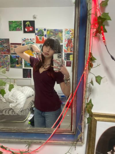 A young woman with long dark hair and bangs poses for a selfie in a mirror. She wears a dark V-neck shirt, jeans, and numerous rings and bracelets. The mirror's frame is decorated with greenery and red LED lights. Behind her in the reflection are colorful paintings hanging on the wall. A bed with white and grey striped sheets is partially visible in the lower right corner. The overall aesthetic is eclectic and artistic.