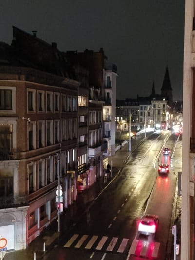 This image captures a nighttime urban street scene on Rue Lambert Lombard in Liège, Belgium, viewed from an elevated perspective. The environment is dark, with artificial lighting originating from streetlights, vehicle lights, and illuminated building interiors, casting reflections on the wet asphalt road surface, indicative of recent precipitation.
In the immediate foreground, a white pedestrian crosswalk is clearly delineated across the wet street. A white sedan with illuminated red brake lights is positioned on this crosswalk, facing away from the viewer. To the left, a multi-story building with a brick facade and rectangular windows dominates the scene's left side. A white circular traffic sign with a red border and horizontal bar, indicating "No Entry," is visible on a pole at the lower left, accompanied by a supplementary panel that states "SAUF CYCLISTES" (except cyclists). A white security camera is mounted on the building's facade.
Further along the wet road, which curves gently to the right, a second dark-colored vehicle with illuminated red taillights is visible, followed by a larger public transport vehicle, identified as a bus by its distinct size, prominent red taillights, and the "BUS" marking painted on the road surface. One individual is discernible on the sidewalk to the left, walking away from the camera.
The street is continuously lined with multi-story buildings, characteristic of European architecture, extending into the midground and background. In the distant background, against the dark sky, the distinctive twin spires of the Basilica of Saint Martin are visible, rising above the city's rooftops. The scene depicts a moderately active urban thoroughfare under nocturnal, damp conditions.