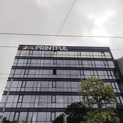 The image captures the Printful office building located on Miera iela in Riga, Latvia. The building is a modern, multi-story structure with a facade of dark-framed, reflective glass windows. Above the windows, the prominent Printful logo is visible, featuring colorful triangular shapes and white lettering. Power lines are strung across the cloudy sky in the upper portion of the image. A tree with green foliage is positioned in front of the building, partially obscuring some of the lower windows. The overall impression is that of a contemporary urban business environment under an overcast sky.