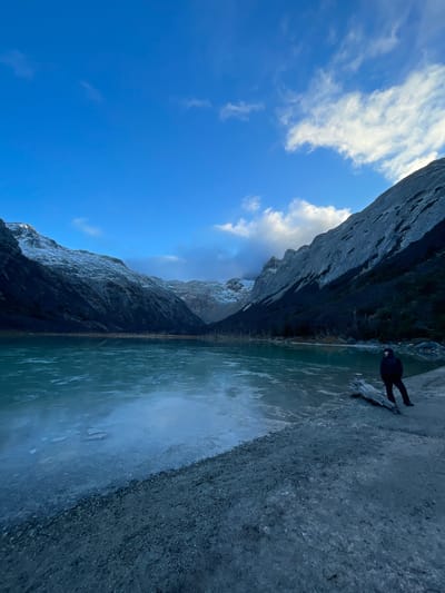 A wide-angle landscape photograph, taken in Unknown, Argentina, shows a partially frozen lake bordered by steep, rugged mountains. The sky is a bright blue with scattered white clouds. The water in the lake is a muted teal color with patches of ice visible on the surface. On the right side of the frame, a person stands on a gravelly shore, facing the lake. They are wearing dark clothing and appear small in relation to the overall scene. The mountains rise sharply on both sides of the lake, their rocky surfaces partially covered in snow. The photograph is composed in a way that emphasizes the scale and grandeur of the natural environment.