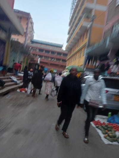 A street scene with pedestrians walking, some blurred due to motion. Vendors display tomatoes and avocados on the ground. Buildings flank the street, varying in height and color from orange to yellow. The sky appears overcast. A car is visible in the background. The overall atmosphere suggests a bustling market area.