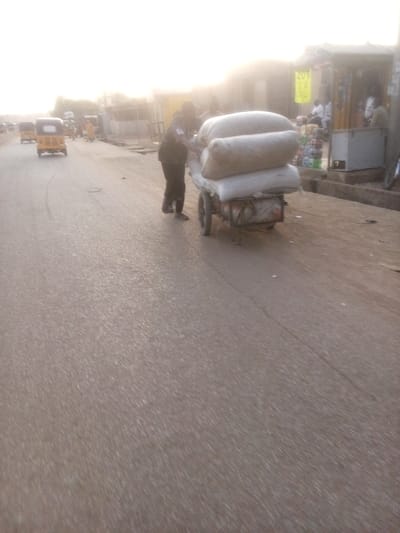 The image captures a vibrant street scene in Rangaza, Nigeria, likely during late afternoon or early morning due to the warm, hazy sunlight filtering through the background.
The central subject is a person, dressed in dark clothing, pushing a two-wheeled handcart laden with three large, light-colored sacks or bales. The cart appears to be moving from left to right along the side of a paved road.
In the mid-ground on the left, several yellow auto-rickshaws, commonly known as Keke Napeps, are visible on the road, suggesting ongoing traffic or a busy thoroughfare. The road surface itself is asphalt, showing some cracks.
On the right side of the frame, alongside a sidewalk, are several informal commercial establishments or market stalls. People are seen near these stalls, some seated. One stall features a small white refrigerator or cooler, and various items are displayed for sale. A yellow sign hanging from an awning over one of the stalls shows blurry text, possibly "20N" or a similar numerical indicator, likely a price or denomination.
The overall setting is characteristic of a bustling urban or semi-urban area in Nigeria, with a mix of street vendors, local transportation, and daily activity under a bright, diffused light. No strong emotions are discernible from the individuals, but the scene conveys a sense of everyday commerce and movement.