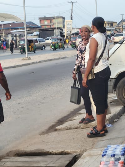 The image shows two women standing on a sidewalk next to a street in Aba, Nigeria. The woman in front wears a patterned shirt, black leggings, and sandals, carrying a black bag. The woman behind her wears a white t-shirt, black pants, and sandals, with a small shoulder bag. The street has auto rickshaws (tuk-tuks) and other vehicles. A sign for "ISICO" is visible in the background. Bottles of water are stacked nearby. The environment appears to be a busy urban commercial area.