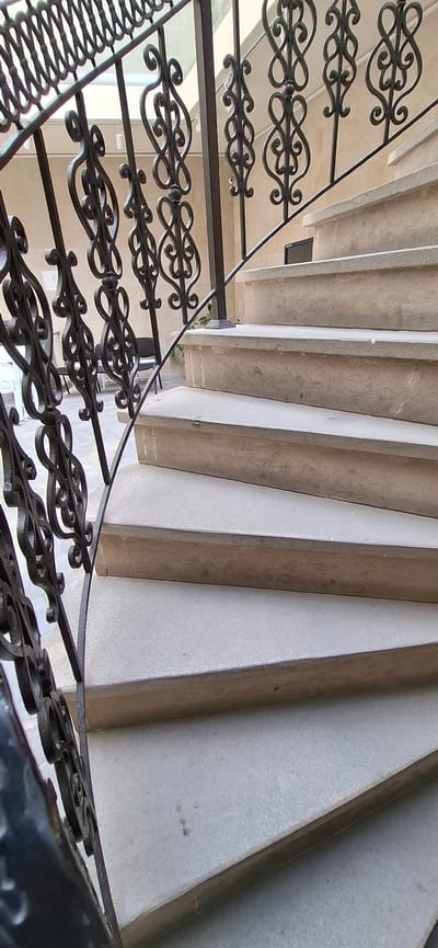 The image shows a close-up, upward-angle view of a stone spiral staircase with a black wrought-iron railing. The staircase is made of light-colored, textured stone steps. The railing features ornate scrolling metalwork. In the background, a textured wall and some chairs are visible. The scene appears to be indoors, possibly in a building with a courtyard or atrium, as suggested by the natural light and the architectural style. There is no clear indication of the specific location in Baku, Azerbaijan.