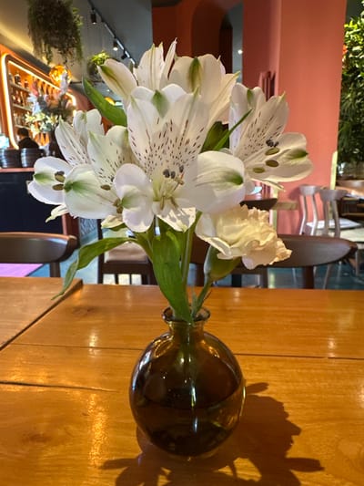A vase containing a bouquet of white lilies and one light yellow carnation sits on a wooden table. The lilies have dark speckles on their petals. The vase is a dark amber color and is partially filled with water. The table appears to be in an indoor setting with a blurred background. The background shows chairs, a bar with bottles on shelves, and hanging plants. There are no visible people in the immediate foreground. The scene appears to be a still life arrangement within a restaurant or bar.