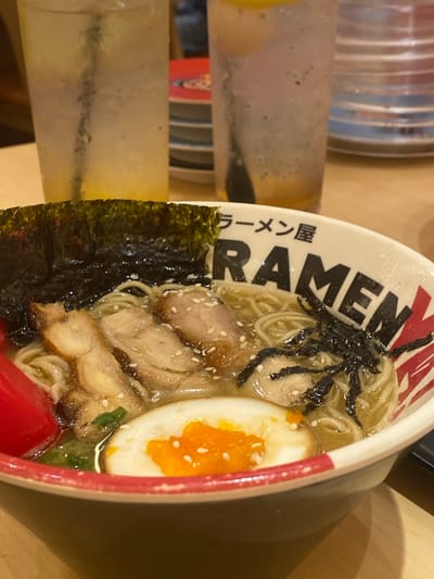 A close-up shot captures a bowl of ramen at a restaurant. The bowl is white with black lettering that spells out "RAMEN" and some Japanese characters above it. The ramen itself is in a clear broth and features noodles, slices of chashu pork, a soft-boiled egg with a bright orange yolk, nori seaweed, and green onions. A red spoon is partially visible on the left side of the bowl. In the background, two glasses of a light-colored beverage, possibly iced tea or lemonade, with ice and straws are visible. The setting appears to be a wooden table in a casual dining establishment. The restaurant is located in Denpasar, Indonesia.