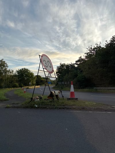 A 30 miles per hour speed limit sign is visible on a metal frame by the side of a road in South Staffordshire, United Kingdom. The sign is circular with a red border and a white background, displaying the number "30" in black. Two traffic cones are placed in front of the sign, one red and white, the other orange. Behind the cones, a collection of objects are piled up, including what appears to be discarded clothing or fabric. The road curves to the left, bordered by grassy verges and trees. The sky is cloudy with hints of sunset.