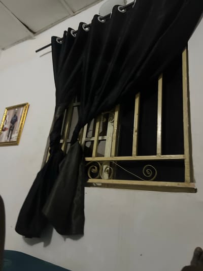 A framed portrait hangs on a white wall, to the left of a barred window covered by a black curtain. The window is ornate, with scrollwork designs on the bars. The room appears to be dimly lit, with the black curtain casting a shadow on the wall. The location is stated to be Howayi, Nigeria.