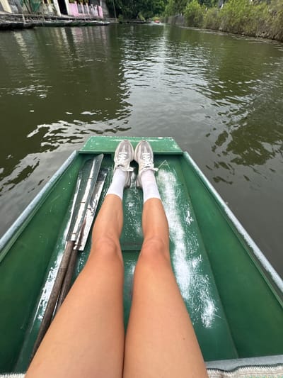 A first-person perspective shot shows a person's legs and feet in a green boat on a body of water. The person is wearing white socks and grey athletic shoes. Two oars with wooden handles and metallic blades are lying inside the boat next to the person's legs. The water is dark green and appears calm, with reflections of the surrounding environment visible on its surface. In the background, a shoreline is lined with buildings and trees. The buildings appear to be of varied colors and architectural styles, with some showing signs of age. The overall scene suggests a leisurely activity, possibly sightseeing or rowing, in a somewhat urbanized natural setting. The location is identified as Nam Hoa Lư Ward, Vietnam.