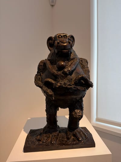 A bronze sculpture of a primate-like figure is displayed on a white pedestal in a well-lit room. The sculpture, which stands upright, is detailed with textured surfaces and appears to be in a gallery setting. The background consists of a light-colored wall and a translucent window blind on the right side of the frame. The lighting is even, casting soft shadows. The location is the Fondation Beyeler in Riehen, near Basel, Switzerland.