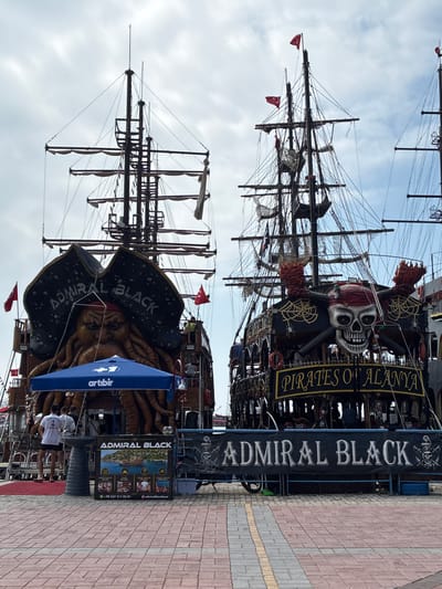 The image shows two pirate-themed boats docked side-by-side in Alanya, Turkey. The boat on the left, named "ADMIRAL BLACK," features a large, ominous kraken-like figure on its stern, wearing a pirate hat. The boat on the right is decorated with a large skull and crossbones, and the text "PIRATES OF ALANYA" is visible on its side. Both boats have multiple masts and rigging, typical of sailing ships. In the foreground, a paved walkway with red and grey bricks is visible. Several people are standing on the dock near the "ADMIRAL BLACK" boat, and a blue umbrella with the "artibir" logo is set up. A banner with "ADMIRAL BLACK" and a smaller image of the boat is displayed on the dock. The sky is overcast with clouds. The overall scene depicts a tourist attraction, likely a pirate boat tour operation in the port of Alanya.