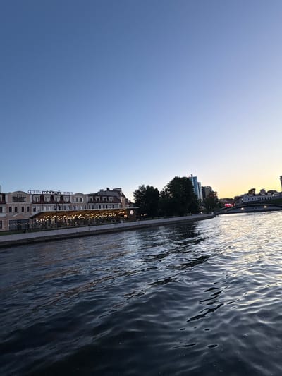 The image depicts a waterside scene at dusk in Minsk, Belarus. The foreground is dominated by rippling water, reflecting the subdued light of the setting sun and the lights from the shore. Beyond the water, a row of buildings lines the embankment. The most prominent of these is a hotel with visible text in Cyrillic that appears to read "ОТЕЛЬ ROBINSON HOTEL". The building is lit with numerous small, warm lights, suggesting it is open for business or dining. Further down the embankment, more modern buildings, including a skyscraper, are visible against the pale twilight sky. Trees are interspersed among the structures, adding a natural element to the urban landscape. The overall mood is tranquil, with the soft colors of the sky and the gentle movement of the water creating a peaceful atmosphere. The time of day is clearly evening, as indicated by the low sun and artificial lighting. The location is identified as Minsk, Belarus. There are no clearly visible people in the image, and the focus is on the cityscape and the water.