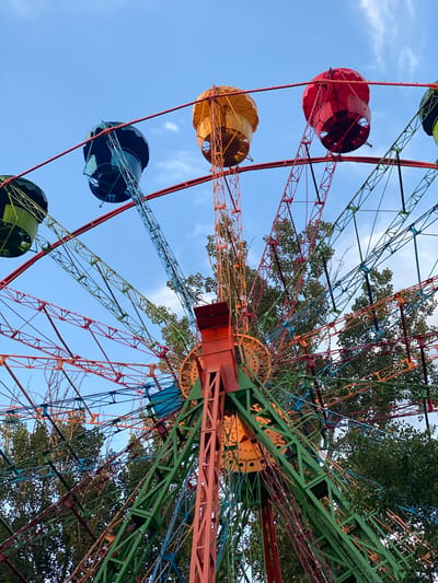 A low-angle, full-frame shot captures a colorful, retro Ferris wheel against a bright blue sky with scattered clouds. The Ferris wheel features vibrant, multi-colored gondolas, including green, dark blue, orange, and red, suspended from a complex network of painted metal struts in red, orange, green, and blue. Tall green trees with dense foliage are visible behind and around the Ferris wheel structure, suggesting an outdoor location. The central support structure of the Ferris wheel is particularly prominent, painted in bold orange and green. This Ferris wheel is located at the First President's Park in Almaty, Kazakhstan. There are no people visible in the image.