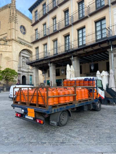 A flatbed truck is parked on a cobblestone street in front of a building with a colonnade in Segovia, Spain. The truck is loaded with numerous orange gas cylinders stacked high, and the truck’s license plate reads “2576 JKY”. The truck has a white cab and a blue chassis. The building behind the truck is a multi-story structure with a large open arcade area. The street is empty except for the truck and a white van parked in the distance. The surrounding environment is urban, with stone buildings and paved roads. The scene is a typical example of the urban environment in the city of Segovia, Spain. The coordinates 40.95, -4.124 place the scene in the Plaza Mayor, a central square in Segovia.