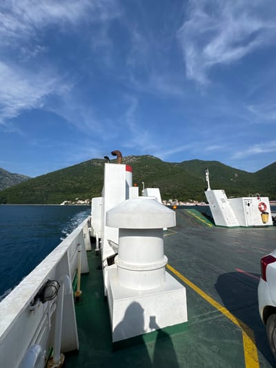 An outdoor, medium-shot, vertical photograph captures a ferry moving through calm, dark blue water, with a heavily vegetated, mountainous coastline in the background. The sky is bright blue with scattered white clouds. In the foreground, the white deck of the ferry dominates the left and center of the frame. A large, white cylindrical structure with a rounded top and a metallic brown chimney with a red band stands on the deck. A white railing runs along the left side of the ferry, with a black light fixture attached. To the right of the center, the ferry's deck is a dark green surface with yellow lane markings. A portion of a white car with a red taillight is visible on the far right. In the distance, the green slopes of mountains rise from the water, dotted with small white buildings and trees. The overall scene suggests a journey through a scenic coastal region, possibly the Bay of Kotor near Prčanj, Montenegro.