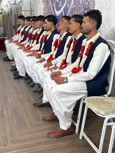 In Quetta City Tehsil, Pakistan, a row of men are seated close together on chairs, likely indoors based on the wooden floor and wall decorations. Each man wears a white shalwar kameez, a navy blue vest, and a garland of red and white flowers. They appear to be part of the same group or event, as their outfits are identical. The men are lined up closely, suggesting a formal gathering or ceremony. The setting appears to be a celebration or special occasion, given the attire of the men and the presence of the garlands.