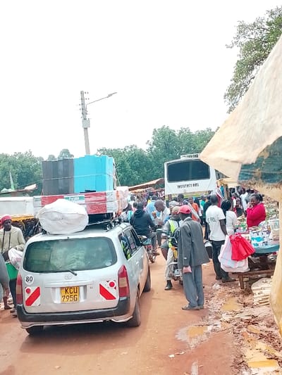 The image shows a busy street scene in Lwandeti, Kenya (latitude: 0.563, longitude: 34.806). A silver Toyota Previa, loaded with goods on its roof rack, is parked on a dirt road with several other vehicles. The car's license plate reads "KCU 7956." There are numerous people walking, shopping, and standing around the car. A white bus with the text "INSTITUTE BUS" visible on its side is parked in the background. Some people are carrying bags and boxes. A few are interacting with vendors at a makeshift market stall with a red and white canopy. The scene is chaotic and colorful, with a mix of people, vehicles, and goods. The road is dusty and covered in muddy puddles. There are trees in the background and a light pole with wires extending from it. The sky is overcast.