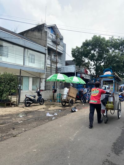 The scene is a street in Bandung, Indonesia, located at coordinates -6.931, 107.602. A man is walking along the street, pushing a food cart. He is wearing a red and yellow shirt with a green design on the back. The food cart is filled with food, and there is a green and white umbrella on the top. Behind him are a few people walking and riding bicycles. In the foreground, there is a black scooter parked on the side of the street. On the left side of the street, there is a building with large windows. There is a green and white striped umbrella in front of the building. The street is paved, and there is some trash on the ground. The sky is gray and overcast. There is a lot of green foliage on the right side of the road.