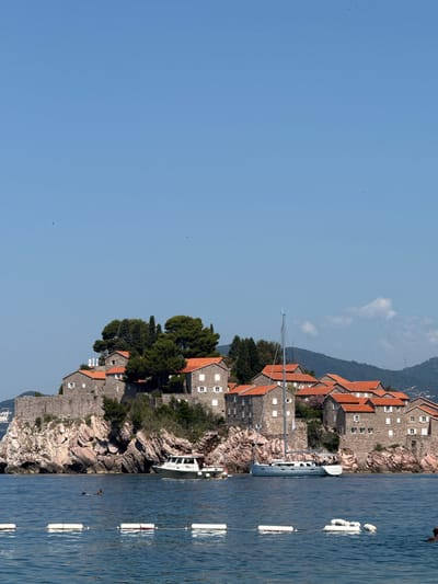 In Pržno, Montenegro, an island settlement composed of stone buildings with red tile roofs is situated on a rocky outcrop. The settlement is surrounded by the clear blue sea, and a sailboat is anchored nearby. A small boat navigates the water close to the shore, and two people are swimming in the sea. The sky above is a clear, cloudless blue, with mountains visible in the distance. The foreground of the image features a rope with buoys, delineating a swimming area.