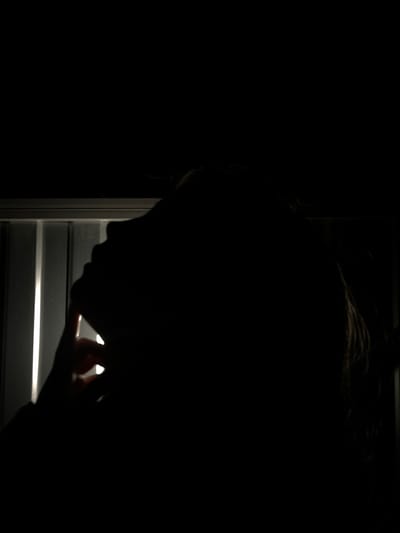 The image captures a deeply shadowed silhouette of a person's head and upper body, viewed in profile. The individual is looking downwards, with the outline of their nose and chin subtly visible against a brighter light source. Their hair appears as a dark, textured mass on top of their head.
The scene is predominantly dark, suggesting an indoor setting during nighttime or in a very dimly lit room. The primary source of illumination emanates from outside, piercing through the gaps of horizontal window blinds or shutters, creating distinct bands of bright light.
One of the person's hands is visible in the lower right, with fingers appearing to rest upon or adjust one of the illuminated slats of the window structure. This action implies the person is actively looking out or observing something beyond the window, or perhaps simply adjusting the blinds.
Notable details include the stark contrast between the intense darkness of the foreground and the luminous horizontal lines, which emphasizes the mysterious silhouette. The time of day is clearly night or a very low-light environment. No specific weather conditions or internal architectural cues are discernible from the image itself that would confirm the location as Novopokrovskaya, Russia. The obscured face and pensive posture evoke a sense of introspection, curiosity, or quiet observation. There is no visible text in the image.