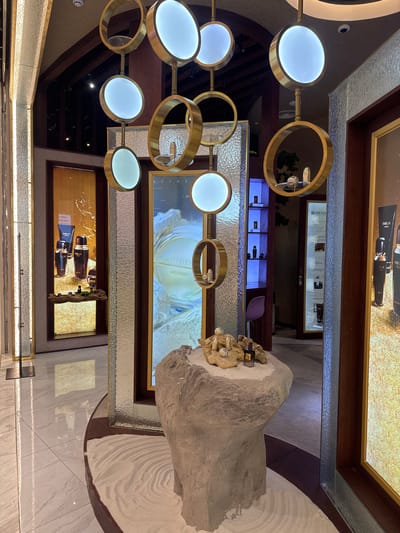 The image depicts the interior of a luxury cosmetics store, likely a Menard boutique, in Hanoi, Vietnam. The store features a minimalist and elegant design with a focus on natural elements and soft lighting. A prominent feature is a large, light-colored stone sculpture serving as a display pedestal, adorned with several small cosmetic bottles and a perfume bottle. Above this, a modern chandelier with circular gold frames and illuminated white discs hangs, casting a warm glow.
The background reveals display cases and promotional screens showcasing Menard skincare products. One screen prominently displays a circular logo with the text "AUTHENTIC MENARD". Another display to the left shows several bottles and tubes of Menard cosmetics against a textured gold background. In the far background, a doorway leads to another section of the store, where shelves with more products are illuminated by blue light. The flooring is a combination of polished white tiles and dark wood accents, with a section of raked white sand in the foreground, adding to the tranquil aesthetic. The overall impression is one of sophistication and a serene shopping experience.