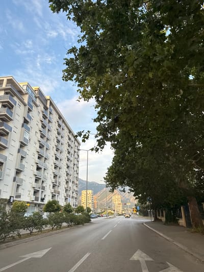 This image depicts a street scene in Bar, Montenegro, likely during the daytime. A large apartment building dominates the foreground, its facade characterized by numerous balconies with dark-colored railings and what appear to be solar panels. The building is a light color, possibly white or beige, and has a modern architectural style.
To the left of the building, a dense canopy of green leaves from a large tree overhangs the scene, creating a natural frame. The sky above is a mix of blue with scattered white clouds.
To the right of the apartment building is a paved road with white lane markings, including a prominent arrow indicating a turn. The road appears to be wet, suggesting recent rain or damp conditions. Several cars are parked along the side of the road, and a line of smaller trees runs between the road and the apartment building.
In the background, across the street, more buildings are visible, including a taller, lighter-colored building with a yellow hue. Mountains can be seen in the distance behind the urban landscape. Streetlights and traffic signals are also present, indicating an active urban environment. The overall impression is a contemporary cityscape with elements of nature, possibly captured in the late afternoon or early morning given the lighting. No people are clearly discernible in the image, nor is there any prominent visible text.