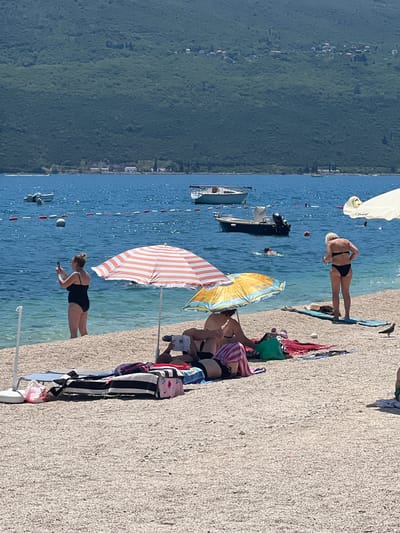 The image depicts a sunny day at a pebbled beach in Baošići, Montenegro. The Adriatic Sea, a deep blue, stretches to the horizon, where a green, tree-covered mountain range forms the backdrop. Several small boats are anchored or moving on the water, and a line of buoys marks a boundary in the sea.
On the beach, several people are enjoying the sun. In the foreground, a woman in a black swimsuit stands at the water's edge, holding a phone to take a picture. Two beach umbrellas, one striped orange and white and the other yellow and orange, provide shade. Under the orange and white umbrella, several people are partially visible, with towels and bags laid out. To the right, a woman in a black bikini stands on the sand. In the middle ground, more people are relaxing on towels, some under umbrellas. A pigeon is visible on the sand to the right of the woman in the black bikini. The ground is covered in small white and grey pebbles.