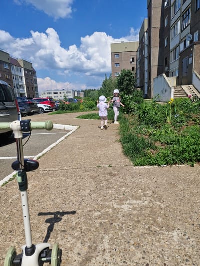 The image depicts an outdoor scene in the city of Марковский, Russia. In the foreground, a light-colored scooter is partially visible, with its handlebars in focus. Two young children, both wearing white hats, are walking away from the camera on a concrete path. The child on the left is wearing a light-colored outfit, while the child on the right is in a grey t-shirt and light-colored pants. Behind the children, there is a garden area with various green plants and some flowers. To the left of the path, a parking lot with several cars is visible, including a dark-colored SUV and a red car. In the background, multi-story apartment buildings dominate the landscape, characterized by their concrete facades and numerous windows. The sky is blue with scattered white clouds. The scene appears to be set on a sunny day.