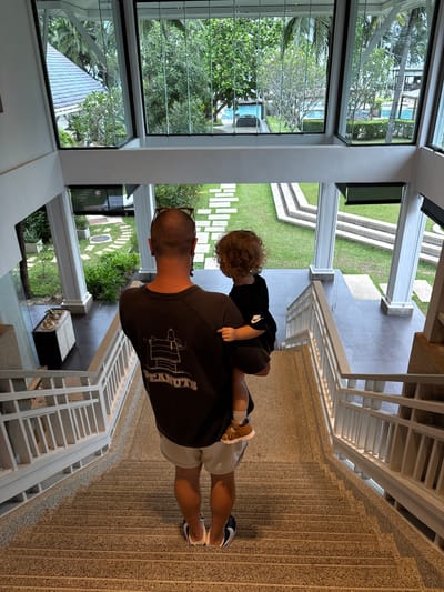 The image depicts a man carrying a young child on his shoulders inside a modern building with large windows overlooking a lush green outdoor space. The man is wearing a dark t-shirt with a graphic print of Snoopy and the word "PEANUTS" visible, and light-colored shorts. The child, with curly blonde hair, is wearing a black t-shirt and brown sandals. They are standing on a textured floor, possibly terrazzo or concrete, with a white staircase to their right. Through the large windows, a swimming pool, palm trees, and manicured gardens are visible, suggesting a tropical location. The scene appears to be indoors, looking out. The lighting suggests it could be daytime. The overall mood is peaceful and familial.