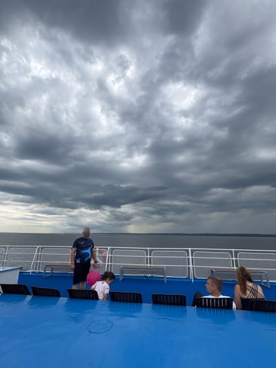 An outdoor scene on a ferry, overlooking a body of water under a cloudy sky. The sky is filled with dramatic, dark clouds, with a hint of rain visible in the distance on the horizon. The water is a deep gray, and the horizon line is marked by a distant shoreline.
In the foreground, the deck of the ferry is painted a vibrant blue. Rows of dark seats are visible, some occupied. On the left, a man wearing a dark patterned t-shirt and black shorts stands looking down, holding a bright pink balloon. Beside him, a young girl in a light-colored top is reaching down, possibly retrieving something from under a seat. Further to the right, a man and a woman are seated, facing away from the camera, looking out at the water. The man is wearing a white t-shirt, and the woman has her hair up and is wearing a dark, strappy top. White railings with netting are visible along the edge of the ferry deck.
The location is Ikskoe Ust'e, Russia. The scene appears to be a moment of travel or observation on the ferry, with the weather contributing to a somewhat somber or dramatic atmosphere.