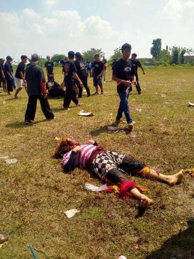 The image depicts an outdoor scene in a dry grassy field within the city of Celep, Indonesia, under a partially cloudy blue sky. In the foreground, a person is lying supine on the ground with eyes closed, dressed in traditional Indonesian attire consisting of a red and white striped long-sleeved shirt, black and white checkered shorts over dark trousers, and red and yellow fabric ornamentation around their shins and ankles. A brown, shaggy headpiece or wig covers their head and face. The person's feet are bare and appear dirty.
Scattered across the dry grass in the foreground and midground are various pieces of litter, including plastic bags, paper plates, and plastic cups. A blue stick-like object is visible in the bottom left corner.
In the midground, a group of approximately 15-20 males, predominantly young adults, are standing and walking across the field. Most are wearing black t-shirts, some with graphic designs, and dark trousers. One individual in blue jeans and a black t-shirt with a patterned sash is walking towards the right, looking towards the viewer. Another individual wearing a black t-shirt with orange visible on the sleeves is walking away from the camera. The group appears to be observing or participating in an event.
The background shows a line of green trees and sparse vegetation, with a light-colored building partially visible through the trees on the right side. The ground throughout the scene is characterized by dry, yellowish-green grass with patches of exposed dirt.