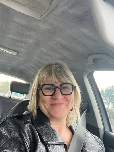 A blonde woman, appearing middle-aged and wearing black-framed octagonal glasses, is seated in the driver's seat of a vehicle in Brignoles, France. She is smiling directly at the camera, secured by a diagonal seatbelt across her chest. She wears a black, textured jacket, possibly leather, over a dark-colored top. A thin necklace and earrings are visible. The vehicle's interior features a light grey, somewhat textured headliner with visible wear or pattern, overhead console elements including a vent, and a passenger-side grab handle. A dark grey headrest from the backseat is visible behind her. Through the windows, daylight is apparent, with trees visible outside the passenger side and a portion of a blue road sign discernible through the rear window, suggesting the vehicle is on a road or highway.