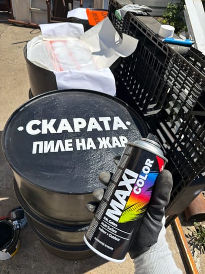 In Gabrovo, Bulgaria, a person wearing black gloves and a grey sleeve is holding a can of black spray paint. The can has colorful text that reads "MAXI COLOR" and "RAL Colors Multi-Surface Indoor + Outdoor Guarantee 10 YEARS". The person is standing near a stack of black barrels. The top barrel has white text painted on it that reads "*СКАРАТА ПИЛЕ НА ЖАР". Part of a black plastic crate with white paper and some orange is visible in the background. The scene appears to be outdoors, with sunlight casting shadows.