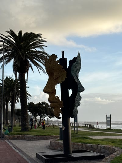 The image shows a metal sculpture of a face, likely located in Cape Town, South Africa. The sculpture is mounted on a black metal pole. The top part of the face is dark metal, while the lower part is a lighter brown color. Below the sculpture, there are palm trees. In the background, there is a boardwalk along a beach, with several people visible. The sky is overcast with grey and white clouds. The ground near the sculpture is paved with bricks and stones.