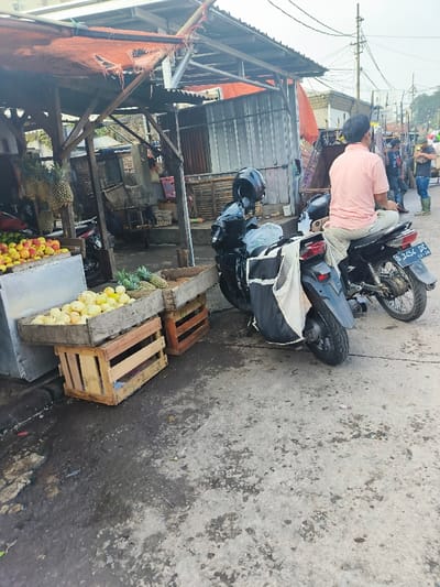 The image shows a street scene in Bandung, Indonesia, located at -6.906 latitude and 107.644 longitude. There are two motorcycles parked in front of a small, open-air market stall. One motorcycle is black, with a black helmet and bag, and the other is white, with a black seat and a black license plate that reads “D 3454 DC”. A man in a pink shirt sits on the white motorcycle, and another man in a blue shirt stands nearby. There are crates of fruit, including pineapples and yellow fruit, on the ground near the stall. The street is paved with gray concrete and there are some wires overhead. The scene appears to be in a busy commercial area.