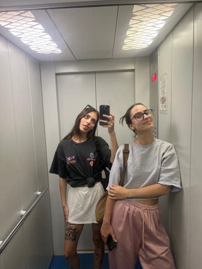 Two young women are taking a selfie in an elevator. The woman on the left is wearing a black t-shirt tied at the waist, a white skirt, and sunglasses on her head. She is sticking her tongue out at the camera. The woman on the right is wearing a grey sweatshirt and pink pants, with glasses and large hoop earrings. Both women appear to be in their late teens or early twenties. The elevator has metal walls and a panel of square lights on the ceiling. There is a red illuminated button on the wall, and a sign with safety instructions. The floor of the elevator is blue. The image does not provide any information about the location of Krasnoyarsk, Russia.