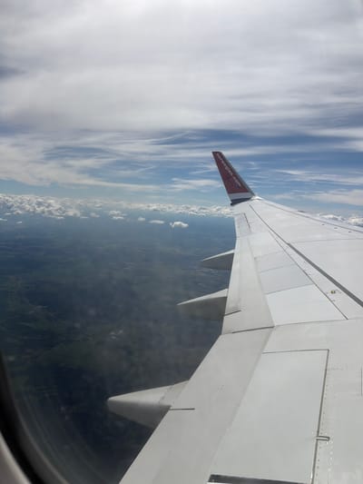 This is an aerial photograph taken from an airplane window, looking out over a landscape. The right side of the image is dominated by the wing of the aircraft, which is white and shows visible panel lines and rivets. The tail fin of the airplane is visible on the upper right, painted red with the text "norwegian.com" in white lettering. Below the wing, a green landscape is visible, with patches of darker green suggesting forests or fields. Roads and some scattered buildings can be discerned within the landscape, indicating a rural or semi-rural area. Above the landscape, clouds are present, ranging from scattered white cumulus clouds to thinner, more wispy cirrus clouds in the distance. The sky transitions from a bright blue at the horizon to a paler blue towards the top of the image, where it is largely obscured by cloud cover. The image does not show any people or specific urban features that would allow for precise identification of Vääna-Jõesuu, Estonia. The perspective suggests the aircraft is in flight at a moderate altitude.
