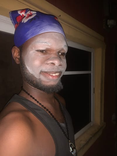 A low-angle, close-up shot captures a person of color, presumably male, indoors in Obosi, Nigeria, at night. The individual's face is largely covered in a light-colored, possibly white, powder or paint, leaving only the dark beard and portions of the skin visible around the eyes and mouth. He wears a blue headscarf adorned with white and red floral patterns. His expression is calm, with a slight smile, and he looks upwards and to the right.
He is wearing a dark, possibly black or grey, tank top and a black beaded necklace with a shiny, ornate pendant hanging on his chest. The background features a window with a white frame, beyond which is darkness, indicating the time of day is night. Around the window, there are wooden structures or frames, likely part of the building's interior, in yellowish and darker brown tones. The lighting in the image is dim, suggesting an indoor setting with limited illumination. There is no visible text in the image.