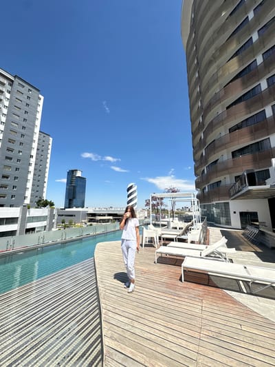 The image captures an elevated outdoor leisure area in Heroica Puebla de Zaragoza, Mexico, under a clear blue sky with minimal white clouds. The foreground consists of a light-colored wooden deck with visible horizontal planks, showing some wear and discoloration. A long, rectangular infinity-edge swimming pool with turquoise water and visible mosaic tiling runs horizontally across the mid-ground.

A single individual, a young adult female, is positioned slightly left of center in the foreground, walking forward and angled slightly towards the viewer. She wears a white t-shirt, light grey sweatpants, and white sneakers with white socks. Her right hand holds a light-colored cup to her mouth, from which she appears to be drinking. Her left hand holds a dark mobile phone.

Beyond the pool and to the right, an outdoor lounge area is visible on an elevated section of the deck. This area features multiple white-framed lounge chairs, some stacked, along with white outdoor tables and chairs arranged under a white pergola structure adorned with red-leafed foliage.

Architectural elements dominate the background. On the far right, a large, curved high-rise building with numerous balconies, characterized by horizontal brown slat-like facade elements, rises vertically and is partially cropped. On the far left, a tall, rectangular grey and white high-rise building with multiple windows is visible. In the distant center, a prominent dark glass skyscraper stands next to a distinctive white and grey spiraled tower or sculpture. The scene depicts a modern, well-maintained recreational space within an urban setting.