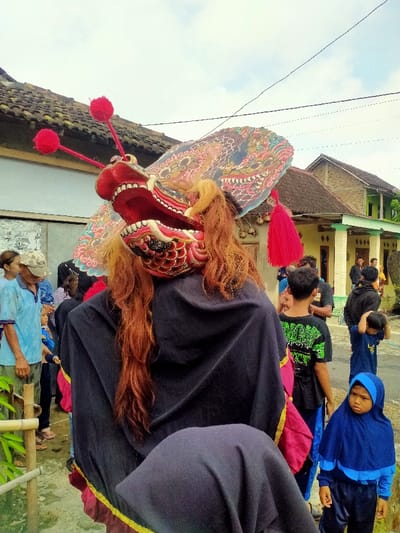 The image shows a person wearing a traditional Indonesian costume with a large, red, painted, wooden mask resembling a dragon's head, adorned with a large, flat, colorful headdress that is decorated with tassels. The mask is open-mouthed with visible teeth, and the wearer's long red hair is flowing over their shoulders. The person is surrounded by a group of people in a street in the village of Kecamatan Kedawung, Indonesia (latitude: -7.514, longitude: 111.03). The person appears to be a performer in a traditional dance. The environment is a residential street with houses and power lines in the background.
