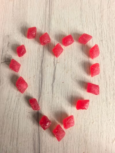 A top-down view shows a heart shape outlined by small, red, diamond-shaped candies on a light-colored, wooden surface. The surface has a visible wood grain pattern running horizontally. There are approximately 17 candies forming the heart shape, with some gaps between them. The lighting creates subtle shadows from the candies onto the surface. There is no indication of a specific location in Sterlitamak, Russia.