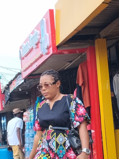 The image shows a woman with braided hair wearing a patterned dress and sunglasses, walking past a building with red and yellow accents. The building has a sign that reads "Cimma Empire". In the background, there's a man wearing a white shirt and tan pants, standing near stacks of water bottles in front of a storefront. The location appears to be a commercial area, possibly a market, in Umuode, Nigeria. Clothing is displayed outside of the building.