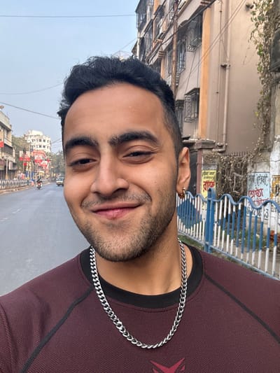 A young man poses outdoors, wearing a burgundy shirt and a silver chain necklace. He has dark hair, a short beard, and a small hoop earring. The background shows a street with buildings and a blue and white barrier. The overall image has a casual, selfie-style feel.