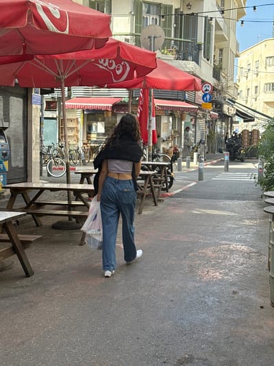 A daytime street-level image depicts an urban commercial area in Tel Aviv-Yafo, Israel. The central figure is a female, viewed from behind, with long, dark, wavy hair. She wears a light purple crop top, loose-fitting blue jeans, and white sneakers, with a dark jacket draped over her shoulders. She is walking away from the camera, carrying a clear plastic bag containing white and red items in her left hand.
The left side of the paved street features several wooden picnic tables with benches, positioned under three large red umbrellas, two of which display the white "Coca-Cola" logo. Behind these, multiple bicycles are parked against a building. A blue public utility box is visible on the far left. The adjacent building is multi-story, light-colored, and features balconies with closed green shutters and a satellite dish.
The street surface is dark gray asphalt with patches of reddish staining. Bollards delineate the path, leading towards a narrow passage where a white-striped crosswalk is visible. A street sign cluster includes a red 'no entry' symbol, a blue pedestrian crossing sign, and Hebrew text. Further into the background, parked motorcycles, indistinct figures, and additional buildings with various shopfronts and awnings are present, beneath overhead string lights.