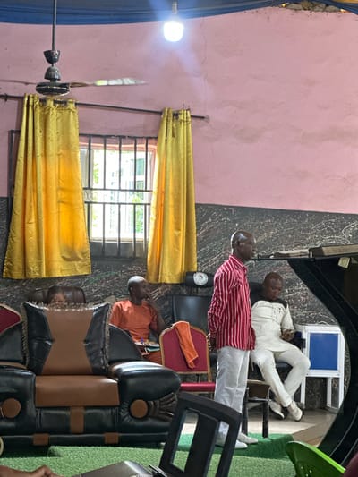 The interior scene shows three men seated in what appears to be a room in Aba, Nigeria. A man in a red and white striped shirt and white trousers sits in the center of the frame, while another man in a white suit and shoes sits further back near what appears to be a staircase. A third man, wearing an orange shirt, is seated in the foreground.
A window with gold curtains is visible, allowing natural light into the room. A ceiling fan with rotating blades is positioned near the window. The room is decorated with patterned walls and includes various pieces of furniture, including chairs with leather cushions. There are objects, such as a bottle and a folded orange cloth, placed around the men. The floor is covered with a mix of materials, including a dark, swirling pattern and artificial green turf.