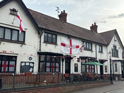 This media file captures the exterior of a building, likely a pub or restaurant, decorated with English flags in anticipation of a sporting event, possibly the World Cup. The building is painted white and has a tiled roof. Various English flags are prominently displayed, including a large one hanging from the facade and smaller ones adorning the windows and strung along the building's eaves.
Notable details include the year "1926" inscribed above one of the windows, suggesting historical significance. Several windows are adorned with large St. George's Cross flags, indicating a strong patriotic sentiment. A blackboard displays information about "WORLD CUP on HERE," confirming the context of a sporting event. Outside, there are tables and chairs, some covered by a green umbrella, suggesting an outdoor seating area. A lamppost stands near the entrance, and bicycles are parked nearby.
The weather appears overcast, and the lighting suggests it is daytime. The overall atmosphere conveys a sense of excitement and national pride. There are also some smaller red and white triangular flags decorating the exterior. Visible text includes "1926" and "WORLD CUP on HERE" on the blackboard, along with other unreadable text on posters and signs. The location is identified as Great Yarmouth, United Kingdom.