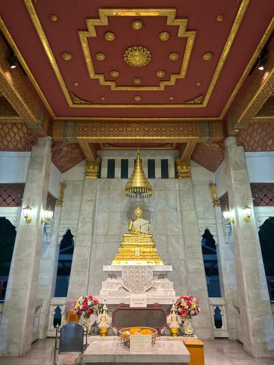 The image depicts a golden Buddha statue seated on a tiered pedestal within a temple-like structure. The statue is adorned with a white cloth draped over its shoulders. Above the statue, a golden tiered umbrella-like structure is suspended. The walls surrounding the statue are made of light-colored marble with ornate golden accents on the pillars and ceiling. The ceiling itself is a deep red with intricate gold patterns and decorative elements. In front of the pedestal, there are two vases filled with colorful flowers, a small statue, and a basket containing marigold flowers. A dark plaque stands to the left of the flowers. The lighting is dim, with lamps mounted on the pillars casting a warm glow. The scene appears to be within a covered area, with openings in the walls revealing glimpses of dark outdoor spaces. The overall impression is one of solemnity and reverence. This scene is likely located in Tha Khlong Town Municipality, Thailand, given the architectural style and religious iconography characteristic of Buddhist temples in the region.