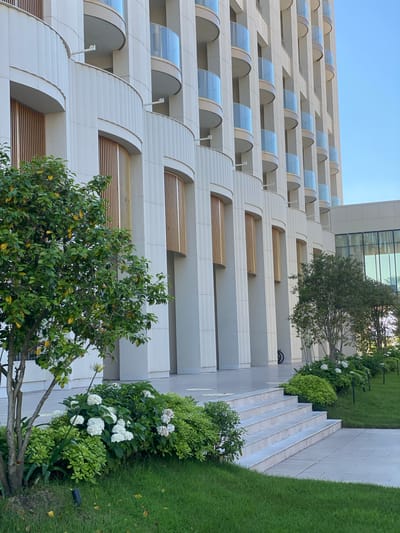 This is a low-angle, eye-level, full exterior shot of a modern, multi-story building with a light-colored facade and repeated curved balconies with glass railings. The building is characterized by vertical fluted columns and wooden slat accents within arched recesses. In the foreground, a well-maintained landscape features a small tree with green leaves, a bush with white flowers, and neatly trimmed green grass. To the right, a set of marble stairs leads up to the building's entrance, flanked by more greenery. The sky is clear and blue. The building is located in Sirius, Russia, specifically in the area of the Sirius Federal Territory, likely a resort or hotel complex. No people are visible in the frame.